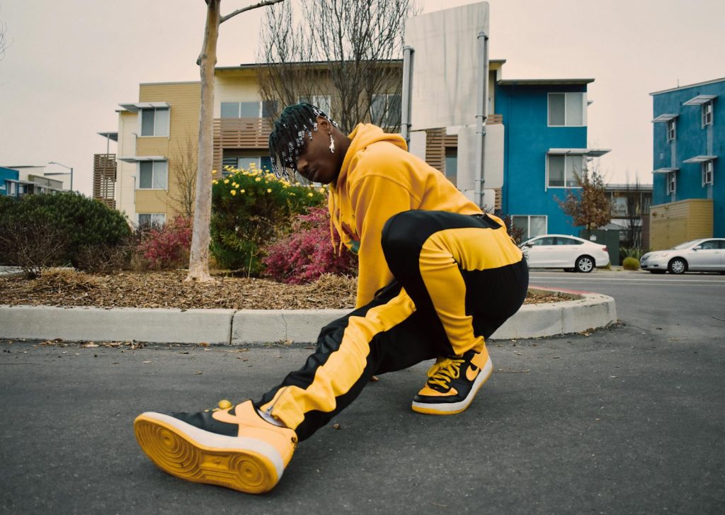 Young African American breakdancer with afro braids in modern yellow black tracksuit doing dance move on asphalt road in residential area